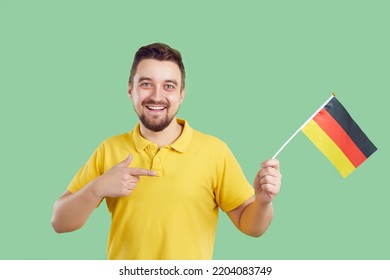 Happy Man Standing Isolated On Green Background, Holding Flag Of Germany And Smiling. Cheerful Handsome Guy Pointing At Flag Of Home Country In His Hand. Young Man Learning German For Job Relocation