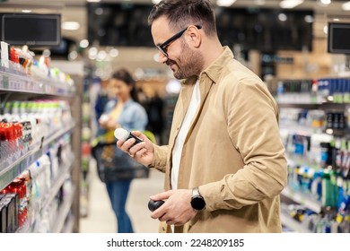 A happy man is standing next to a shelves in with deodorants in supermarket and choosing one.