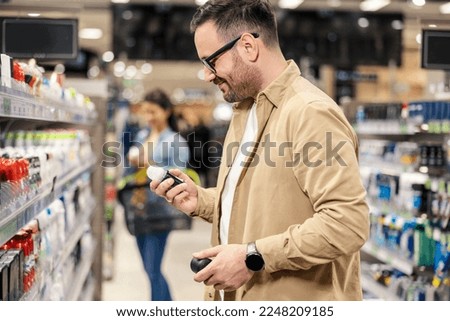 A happy man is standing next to a shelves in with deodorants in supermarket and choosing one.