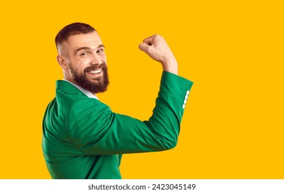 Happy man in suit shows his strong biceps muscles. Cheerful handsome fit bearded young guy in green jacket flexes his arm, raises fist, looks at camera and smiles. Studio portrait, side profile view