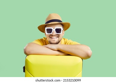 Happy man with suitcase who is waiting for summer trip isolated on light green background. Portrait of young smiling man in sunglasses and summer hat leans on yellow suitcase and looks at camera. - Shutterstock ID 2153210099