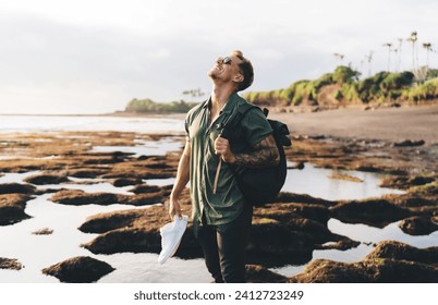 Happy man in sunglasses and casual clothes with backpack looking up while standing alone near sea shore against picturesque scenery of blurred background - Powered by Shutterstock