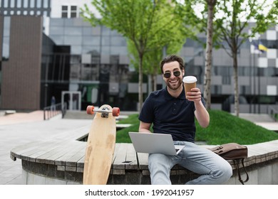 happy man in sunglasses and polo shirt holding paper cup and using laptop near smartphone and longboard - Powered by Shutterstock