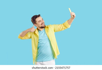 Happy Man Takes Selfie On Cellphone. Cheerful Young Guy In Blue T Shirt And Yellow Shirt Standing Isolated On Blue Background, Smiling, Giving Thumbs Up And Taking Photo Of Himself On His Mobile Phone