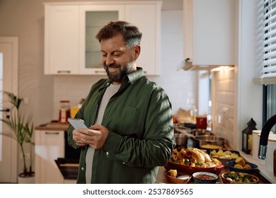 Happy man texting on mobile phone after cooking for Thanksgiving meal in the kitchen.  - Powered by Shutterstock