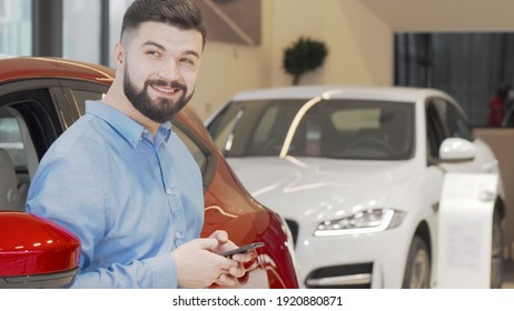 Happy Man Texting On A Smart Phone While Shopping For New Car At The Dealership. Cheerful Young Man Using His Mobile Phone At Automobile Dealership Salon. 4k Footage