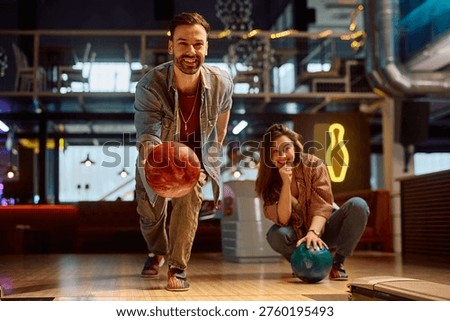Happy man throwing a ball while playing with his girlfriend at bowling alley. 