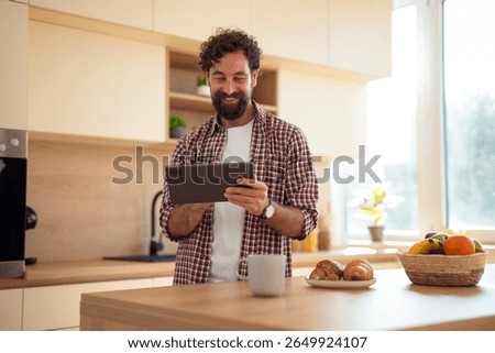 Happy man using a digital tablet in a sunlit kitchen, standing by a counter with coffee, croissants, and a bowl of fruit.