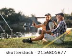 Happy man using smart phone and laughing with his senior father while fishing and camping in nature. 