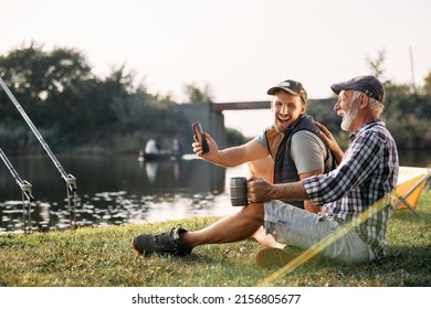 Happy man using smart phone and laughing with his senior father while fishing and camping in nature.  - Shutterstock ID 2156805677