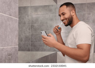 Happy man using smartphone while brushing teeth in bathroom, space for text. Internet addiction