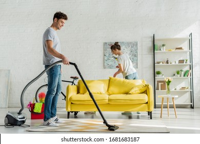 Happy Man Using Vacuum Cleaner Near Woman In Living Room