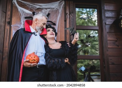 Happy Man In Vampire Costume Holding Carved Pumpkin And Embracing Wife On Cottage Porch With Halloween Decoration