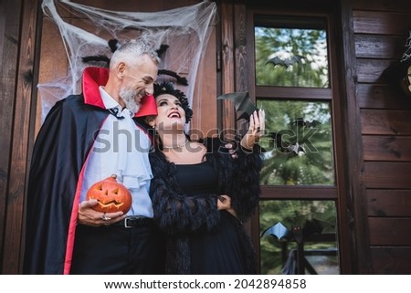 happy man in vampire costume holding carved pumpkin and embracing wife on cottage porch with halloween decoration