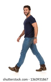 Happy Man Is Walking In Jeans, Boots And Blue T-shirt And Looking At Camera. Side View. Full Length Studio Shot Isolated On White.