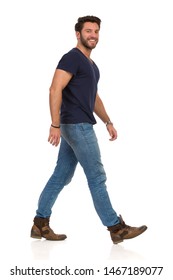 Happy Man Is Walking In Jeans, Boots And Blue T-shirt And Looking At Camera. Side View. Full Length Studio Shot Isolated On White.