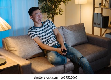 Happy man watch tv in evening at home living room. young asian korean male sitting on sofa and laughing cheerful while watching funny movie on television in late night. joyful guy enjoy leisure time