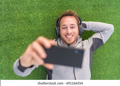 Happy Man Watching Phone Video App Listening With Headphones And Smartphone Relaxing Outside On Summer Green Grass.