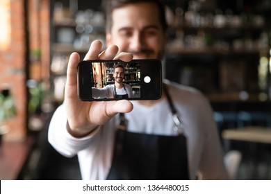 Happy man wearing apron holds smartphone with camera on, waiter or cafe owner make selfie photo focus on device with self portrait image on mobile screen, share life in social media having fun concept - Powered by Shutterstock