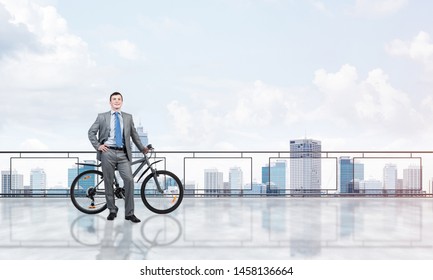 Happy man wearing business suit standing on balcony with bike. Businessman with bicycle on background of sky above megalopolis. Smiling cyclist holding bicycle on terrace with modern downtown view.