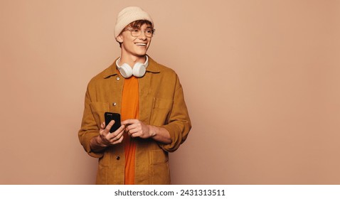 Happy man, wearing casual denim clothing, stands against a peach background in a studio. He holds a smartphone, using a mobile app and texting. Vibrant and colorful, intellectual.