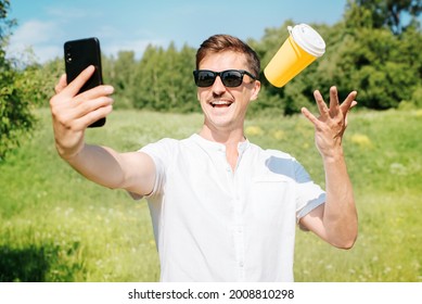 Happy Man Wearing Glasses Tossing Disposable Cup Of Coffee And Taking Selfie On Mobile Phone, Outdoors Summer Day. Portrait Of Smiling Mustachioed Young Man Rejoicing At Phone Camera Outside In Park