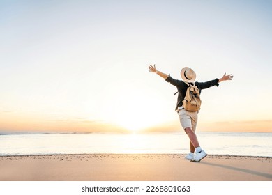 Happy man wearing hat and backpack raising arms up on the beach at sunset - Delightful man enjoying peaceful moment walking outdoors -  Wellness, healthcare, traveling and mental health concept - Shutterstock ID 2268801063