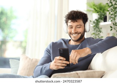 Happy man wearing headphone listening music using phone sitting on a couch at home - Powered by Shutterstock