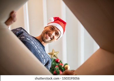 Happy Man Wearing Xmas Hat Unpacking Christmas Gift. Young Man Smiling While Opening Present And Looking Inside. Portrait Of Guy Opening Cartboard Box Under The Christmas Tree While Looking At Camera.