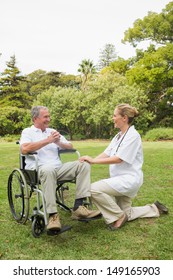 Happy Man In A Wheelchair Talking With His Nurse Kneeling Beside Him On Sunny Day In The Park