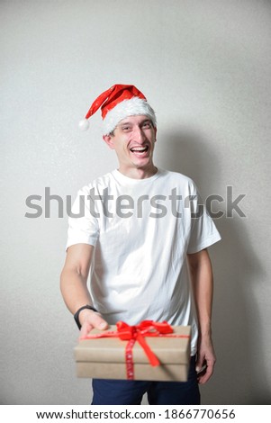 happy man in a white T-shirt and Santa's red Christmas hat holds a gift with a red ribbon on hands. congratulations on christmas. holiday concept in pandemic mode. gift delivery to people.