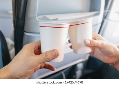 Happy Man And A Woman Clink Paper Cups In The Interior Of An Airplane During A Vacation Flight. Catering And Wine Tasting