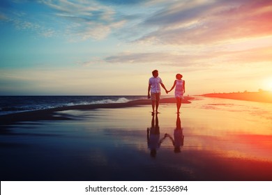 Happy man and woman couple walking and holding hands on a beach with bright colorful sunrise on the background, romantic couple in love walking at sunset, woman and man in love walking hand in hand
