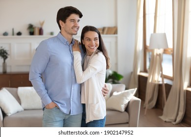 Happy man and woman hugging, standing in modern living room at home, excited young couple looking to aside, smiling wife and husband dreaming, planning good future, visualizing, enjoying leisure time