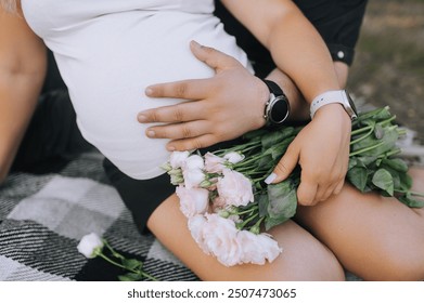 Happy man and woman, mother and father tenderly hug pregnant belly with their hands, sitting on a blanket with flowers outdoors. Close-up photo, pregnancy, motherhood. - Powered by Shutterstock