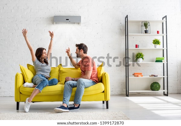 happy man and woman talking while\
sitting on yellow sofa under air conditioner at\
home