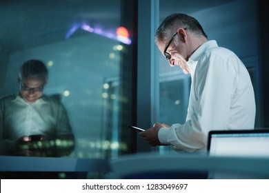 Happy Man Working Overtime In Modern Office Late At Night. Mature Businessman Typing On Cell Phone, With City Lights In Background From Office Window.