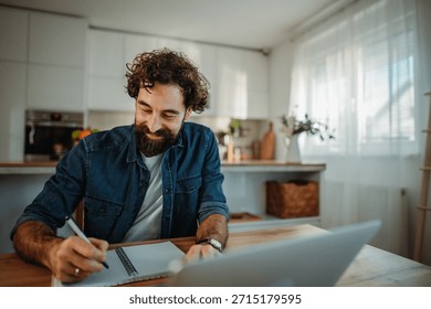 Happy man writes in notebook while working remotely on laptop in bright kitchen
