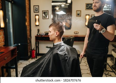 Happy Manly Professional Hairdresser Spraying Client's Hair In A Barber Shop. Letting Drops Fall From Above. He's Standing Behind Client In A Chair Sitting In Profile.