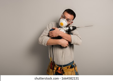 Happy Manual Worker In Safety Mask Hugging Electric Drill On Grey