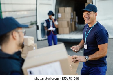 Happy manual worker using touchpad while communicating with his coworker and organizing package delivery. 