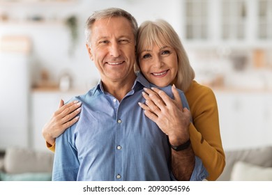 Happy Marriage. Senior Wife Embracing Her Husband Posing Together Holding Hands Smiling Looking At Camera Standing In Kitchen At Home. Retirement Lifestyle Concept