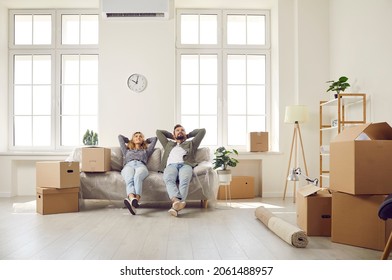 Happy Married Couple Buy New Home And Move In First Own House. Tired Husband And Wife Take Break Resting And Relaxing On Sofa In Spacious Room With High Ceiling And Large Windows. Real Estate Concept