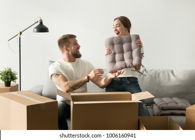 Happy Married Couple Having Fun While Packing In Modern Living Room On Moving Day, Woman Holding Cushion Cheerful Man Helping To Unpack Cardboard Boxes On Couch, Settle Move Into New Home Relocation