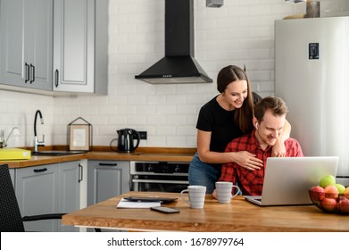 A Happy Married Couple At The Kitchen In A New Home. Attractive Young Man Sits With Laptop, The Beautiful Woman Stands Behind And Hug Him, They Do Online Shopping Together And Smile