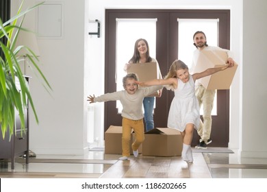 Happy Married Couple And Little Children Arrive At New Modern House. Husband And Wife With Cardboard Boxes Look At Running Inside Excited Daughter And Son. Buy Realestate, Mortgage And Moving Concept