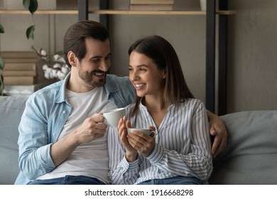 Happy Married Couple Relax On Cozy Sofa At Home Cuddle Discuss Good News Drink Aromatic Tea Strong Coffee. Smiling Young Man Woman In Love Meet Morning Together Sit On Couch Hold Cups With Hot Drinks