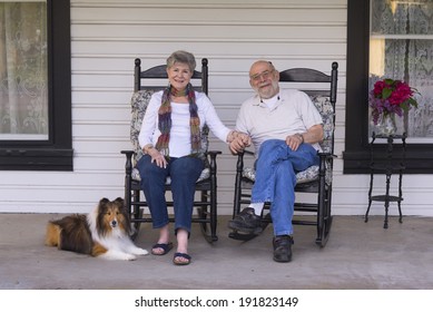 A Happy Married Couple Relax On Their Porch In Matching Rocking Chairs With Their Shetland Sheepdog By Their Side.