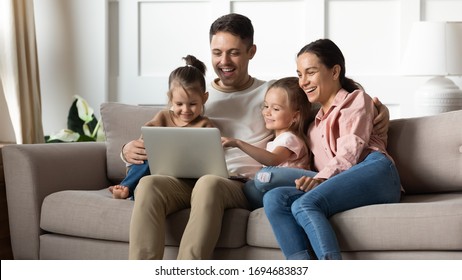 Happy married couple resting on sofa with adorable little children daughters, watching funny movies on computer at home. Laughing family playing online game or shopping together in living room. - Shutterstock ID 1694683837