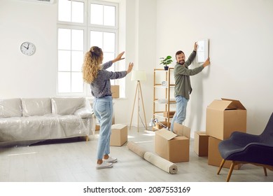 Happy married couple who recently bought house are unpacking stuff and decorating new home. Young man and woman are making living room cozy and choosing place on wall to hang painting or family photo - Shutterstock ID 2087553169